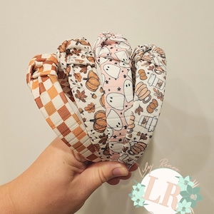 May include: Four different patterned headbands in a hand. The headbands are brown, orange, and white with a checkered pattern, a pumpkin and leaf pattern, a ghost pattern, and a pumpkin and coffee cup pattern. The headbands are tied in a knot.