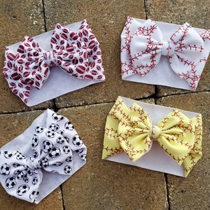 May include: Four white headbands with different sports-themed fabric bows. The bows are made of a soft, stretchy fabric and feature a variety of sports-themed prints, including footballs, baseballs, and soccer balls.