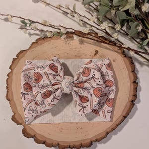 May include: A baseball-themed baby headband with a large bow. The bow features a repeating pattern of baseball bats, gloves, and bows in shades of orange and white on a light pink background. The headband is on a wooden slice.