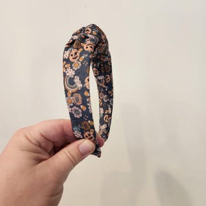 May include: A dark blue headband with a knotted design. The headband is covered in a print of orange pumpkins, candy corn, and flowers.