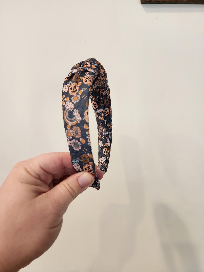 May include: A dark blue headband with a knotted design. The headband is covered in a print of orange pumpkins, candy corn, and flowers.