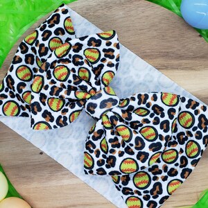 May include: A white headband with a large bow featuring a softball and leopard print design. The bow has a repeating pattern of yellow softballs with red stitching and a brown leopard print on a white background. The headband is on a wooden surface with Easter eggs.
