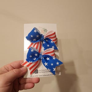 May include: Two red, white, and blue American flag hair bows. The bows are made of ribbon and have a white center. The bows are attached to a white card with the text "Lily Ranee Bows Lily Ranee Boutique Visit us online: www.LilyRaneeBoutique.com".