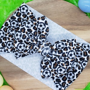 Puede incluir: Un lazo para el pelo de color marrón y blanco con un diseño de balón de fútbol. El lazo está sobre una superficie de madera.