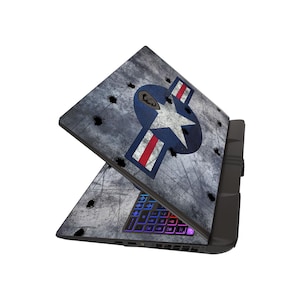 May include: A laptop with a distressed gray finish and a military-themed design. The design features a blue emblem with a white star and red stripes, along with simulated bullet holes. The keyboard has a multi-colored backlight.