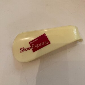 May include: A cream-colored shoe horn with the text "Shoe Express" in red on a red rectangle. The shoe horn is curved and has a smooth surface, designed to assist in putting on shoes.