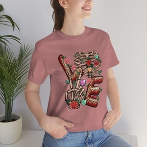 Puede incluir: Una camiseta rosa con un gráfico de una mano de esqueleto haciendo un signo de paz con las letras "LOVE" en un diseño de estampado de leopardo. Las letras están decoradas con rosas y corazones de caramelo.
