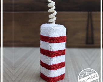 May include: A red and white striped firecracker with a white curled wick on top. The firecracker is made with a punch needle technique and is a handmade craft.