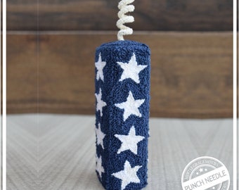May include: A blue and white fabric firecracker with white stars. The firecracker is decorated with a white, curly, paper-like fuse. The firecracker is sitting on a light brown surface. The image includes a white circle with the text "Myrtle Grace Motifs Punch Needle Myrtle Grace Motifs".