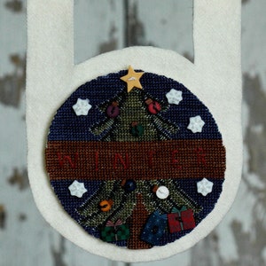 May include: A white fabric Christmas wall hanging with a wooden rod. The circular design features a cross-stitched Christmas tree, the word "WINTER", and decorative elements like stars and ornaments. The hanging measures approximately 10 inches in width.