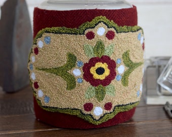 May include: A clear glass jar with a wooden lid. The jar is wrapped in a burgundy fabric with an embroidered tan patch featuring a floral design in red, green, yellow, and blue. The jar is a decorative storage container.