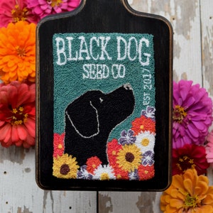 May include: A black wooden chopping board with a green and white needlepoint design. The design features a black dog with a floral border and the text "Black Dog Seed Co. Est. 2017".
