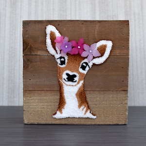 May include: A brown wooden plaque with a white and brown felt deer head with a pink and purple flower crown.
