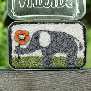 Peut inclure: Un éléphant gris avec une défense blanche et une oreille blanche tient une fleur orange vif. L'image est dans une boîte en métal avec le mot "Vinolde" sur le couvercle.
