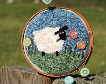 May include: A white sheep with black eyes and a pink flower in its mouth is embroidered on a blue background. The embroidery is in a wooden hoop with a brown frame. There are colorful buttons around the hoop.