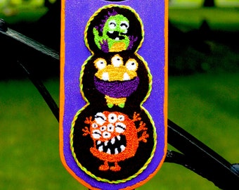 May include: A purple door hanger with an orange border. The hanger features three embroidered monsters in a stack. The top monster is green with a yellow mouth and eyes. The middle monster is purple with yellow eyes and a yellow mouth. The bottom monster is orange with a black mouth and many eyes.