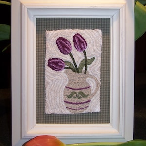 May include: A white framed picture with a purple and green embroidered image of three purple tulips in a brown and white pitcher on a green and white gingham background.