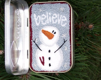 May include: A small, silver tin with a white snowman and the word "believe" on a blue background. The tin is decorated with a red border.