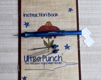May include: A blue Ultra Punch needle tool with a spring and a set of needles. The tool is on a tan instruction booklet with the text "Instruction Book" and "Ultra Punch The Precision Adjustable Needle!" The booklet also has a red flower design and the website address "www.ultrapunchneedle.com".