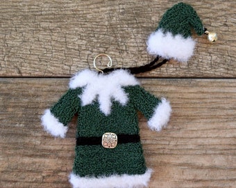 May include: A green elf costume ornament with a white fur trim and a black belt with a silver buckle. The ornament also includes a matching green elf hat with a white fur trim and a silver bell.