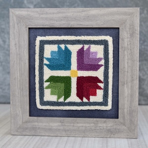 Peut inclure: Un motif de point d'aiguille encadré présentant un motif géométrique coloré avec quatre sections de bleu, violet, vert et rouge. Le motif est sur un fond blanc et encadré dans un cadre en bois gris.