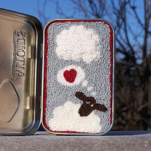 May include: A small tin with a red border, containing a gray felt background with a white cloud, a red heart, and a brown sheep. The sheep is in a white cloud and has a thought bubble above its head.