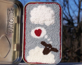 May include: A small tin with a red border, containing a gray felt background with a white cloud, a red heart, and a brown sheep. The sheep is in a white cloud and has a thought bubble above its head.