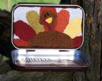 May include: A small metal tin with a brown, orange, yellow, and white needlepoint design of a turkey. The tin is open and the inside is silver with the word "ANTONIA" printed on the bottom.