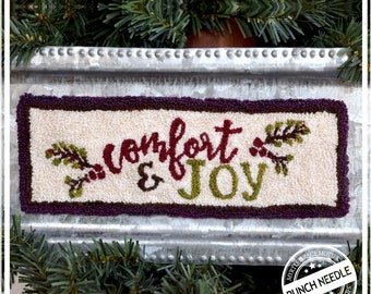 May include: A white needlepoint canvas with a purple border and the words "comfort & joy" in red and green lettering. There are two small green sprigs with red berries on either side of the text.
