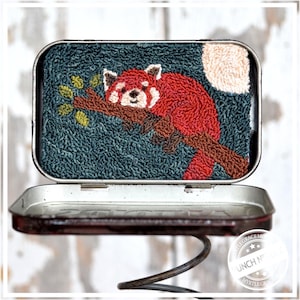 May include: A red panda is depicted in a needlepoint design on a dark blue background. The red panda is curled up on a branch with a full moon in the background. The design is framed in a metal tin.