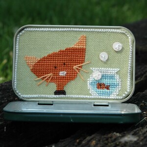 May include: A green and white tin with a cross-stitch design of an orange cat looking at a fishbowl with a small orange fish inside. The tin has two white buttons on the top.