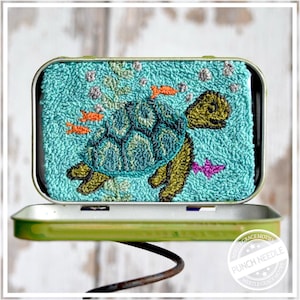 May include: A turquoise punch needle art piece featuring a sea turtle swimming among orange fish and bubbles. The artwork is displayed inside a green metal tin. The turtle has a green and blue shell and is surrounded by a turquoise background.