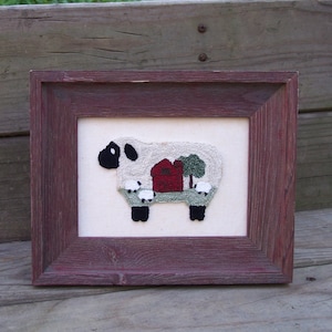 May include: A framed needlepoint picture of a white sheep with a red barn and green trees in the background. The sheep is standing on a green field.