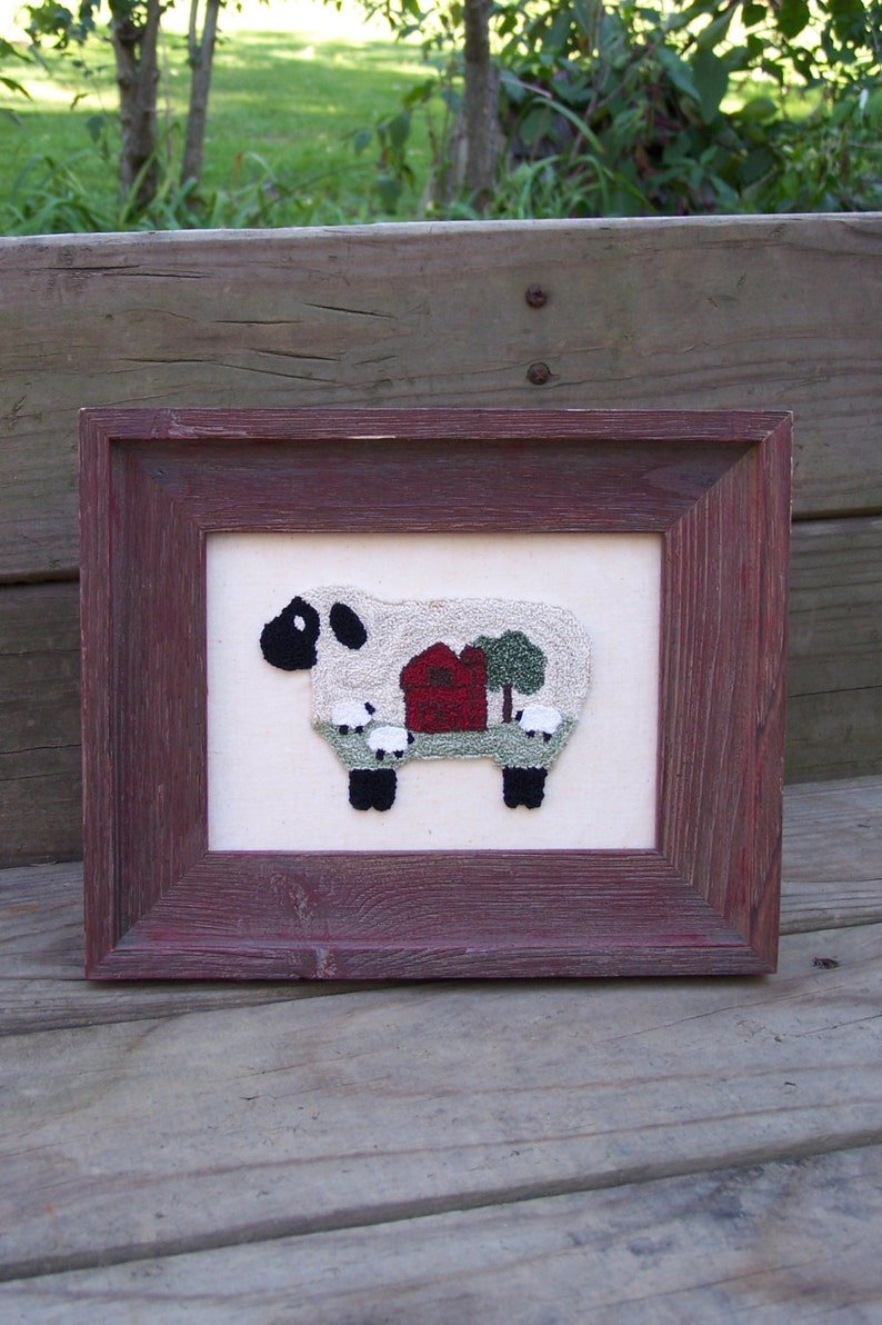 May include: A framed needlepoint picture of a white sheep with a red barn and green trees in the background. The sheep is standing on a green field.