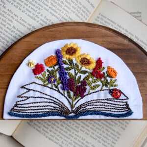 May include: Embroidered bookmark featuring an open book with colorful flowers and a ladybug. The flowers include sunflowers, orange blooms, purple sprigs, and red flowers. A small yellow bee is also embroidered on the white fabric.