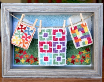 May include: Three colorful miniature quilted squares hang on a clothesline in a gray frame. The squares are made of yarn and feature geometric patterns in shades of red, orange, yellow, green, blue, and purple.