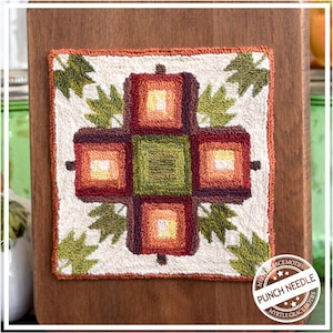 Könnte beinhalten: Ein quadratisches, cremefarbenes Punch-Needle-Kunstwerk mit einem zentralen Kreuzdesign in Orange-, Rot- und Grüntönen. Grüne Blätter sind in jeder Ecke platziert. Das Stück ist auf einer Holzplatte montiert. Der Text "PUNCH NEEDLE" ist sichtbar.