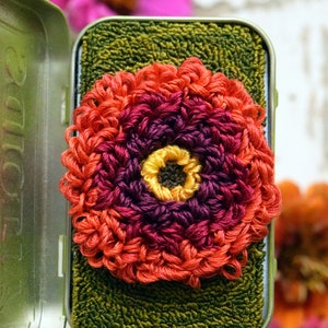 Op de afbeelding: Een handgemaakte, kleurrijke gehaakte bloem in tinten oranje, rood, paars en geel, in een groene metalen doos. De bloem is gecentreerd op een groene, getextureerde ondergrond. De achtergrond bevat vage roze en oranje bloemen.