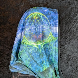 Può includere: Una sciarpa con cappuccio con un vivace design psichedelico. Il tessuto presenta un motivo blu e verde con un design circolare centrale. La sciarpa è drappeggiata su uno sfondo scuro, mettendo in risalto la stampa colorata.