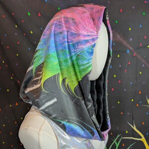 Può includere: Un mantello con cappuccio con un design astratto vibrante. Il cappuccio presenta un gradiente di rosa, blu, verde e giallo, con uno sfondo nero e grigio. L'interno è foderato in tessuto nero. Il mantello è esposto su un manichino.