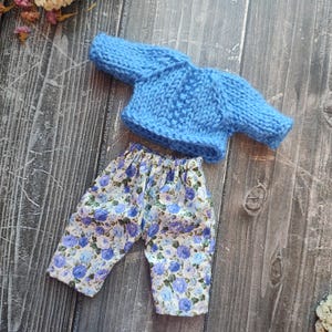 May include: A blue knitted sweater and floral print pants for a doll. The sweater has a cable knit pattern and the pants have a white background with blue flowers.