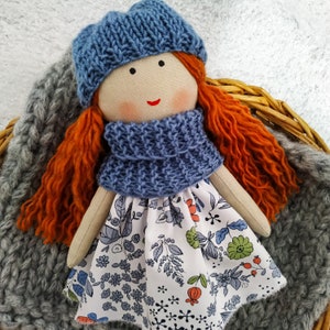 May include: A handmade rag doll with a blue knitted hat and scarf, a white dress with floral print, and long red hair.