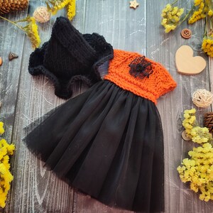 Peut inclure: Un chapeau pointu en tricot noir et une robe avec un corsage en tricot orange orné d'une fleur en dentelle noire et une jupe en tulle noir. L'ensemble est conçu pour une petite poupée ou figurine, mettant en valeur des textures contrastées.