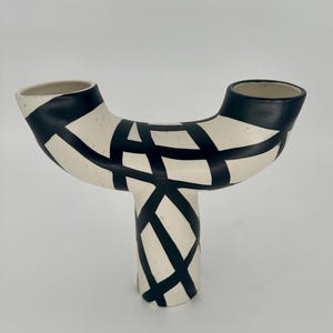 May include: A ceramic vase with a unique, abstract design. The vase is off-white with a black geometric pattern. It has a T-shape with two openings at the top and a single base. The design is modern and artistic.