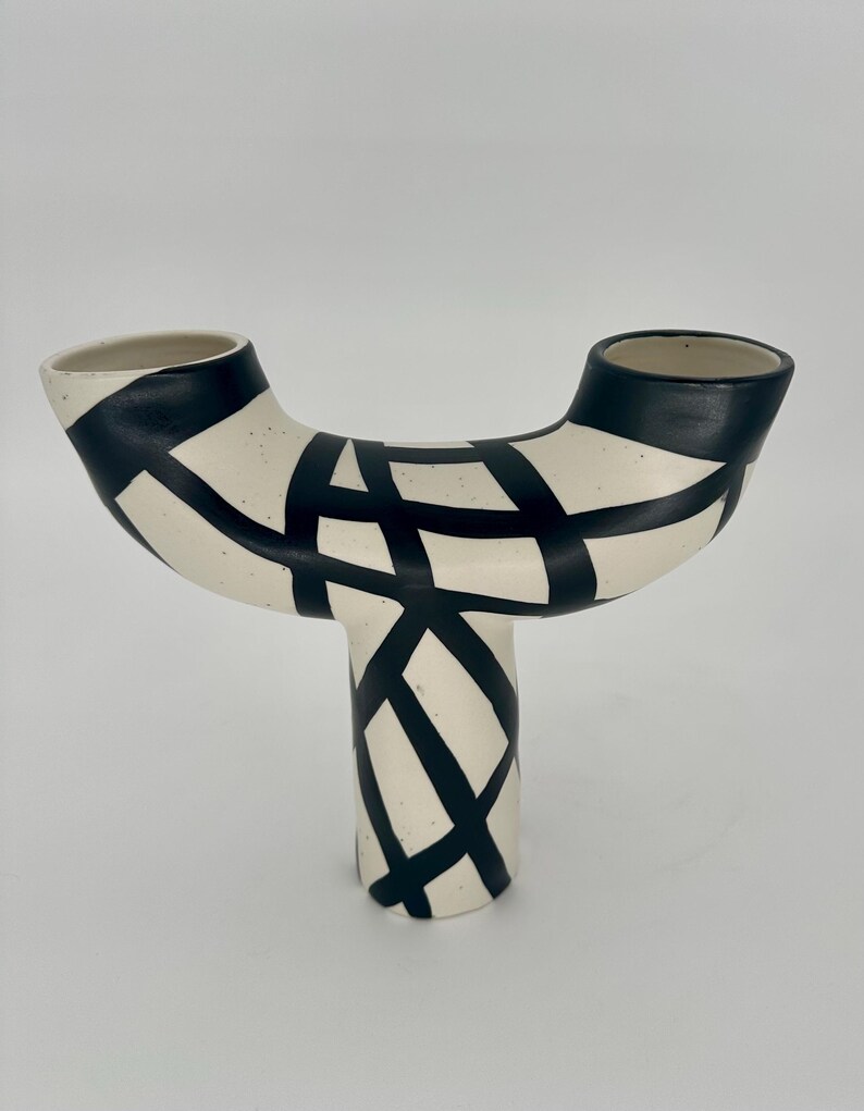 May include: A ceramic vase with a unique, abstract design. The vase is off-white with a black geometric pattern. It has a T-shape with two openings at the top and a single base. The design is modern and artistic.