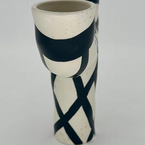 May include: A pair of ceramic vases with a cream-colored base and black geometric designs. One vase is taller and cylindrical, while the other is shorter and angled. The vases have a speckled texture.