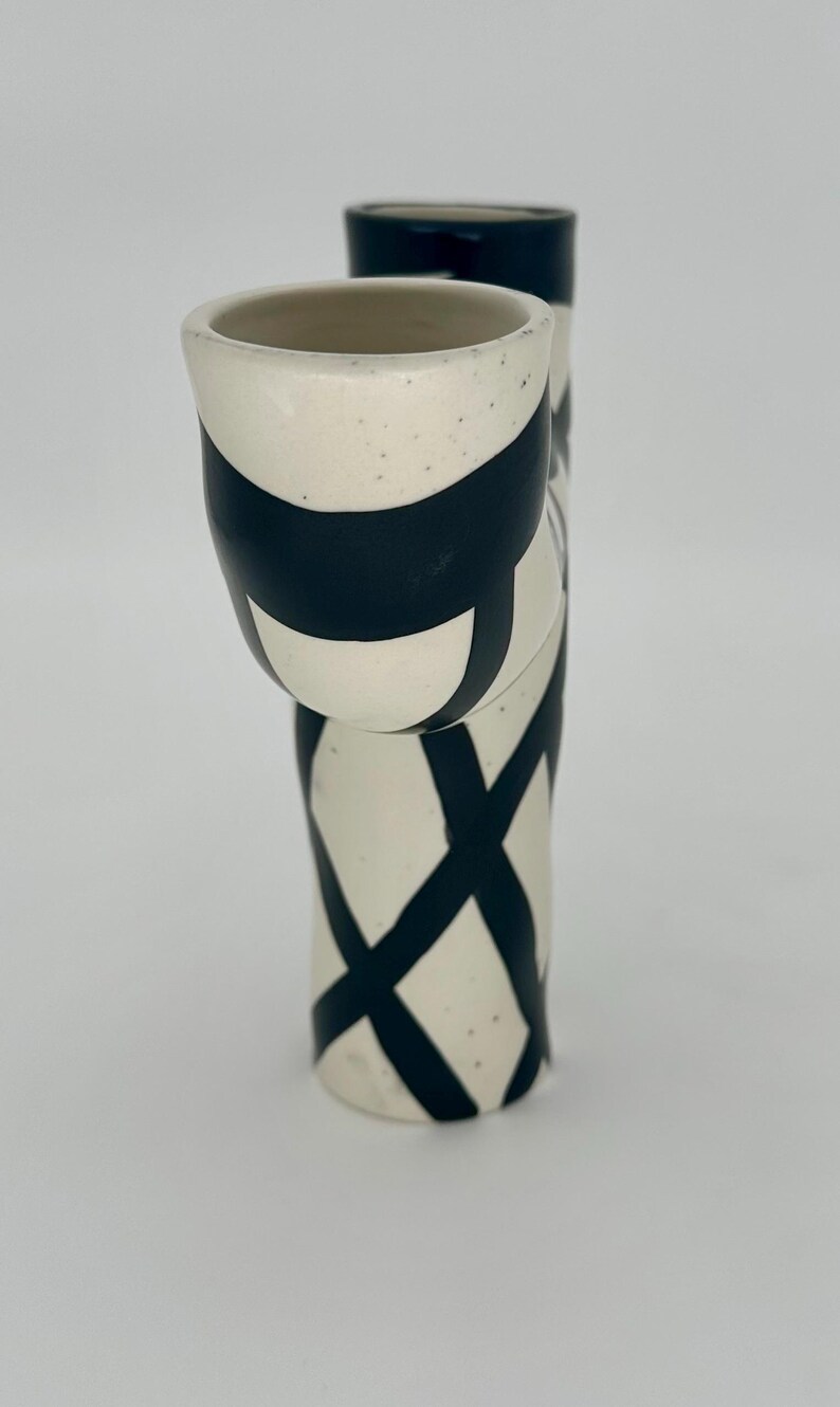 May include: A pair of ceramic vases with a cream-colored base and black geometric designs. One vase is taller and cylindrical, while the other is shorter and angled. The vases have a speckled texture.