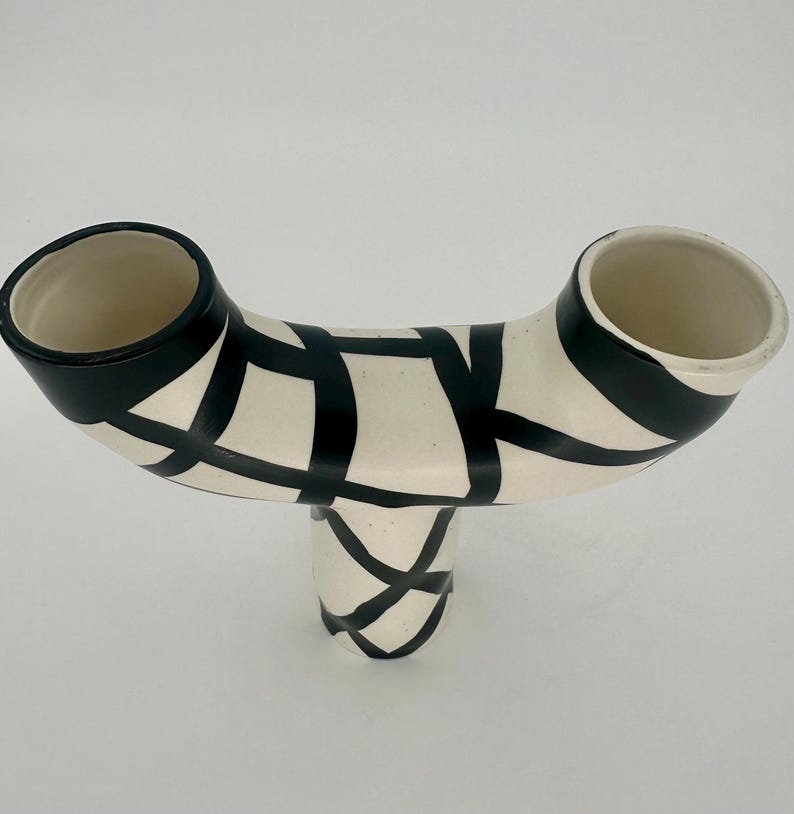 May include: A cream-colored ceramic vase with a unique, curved design. The vase features two openings and a base, all adorned with a black geometric pattern. The rims of the openings are also black.