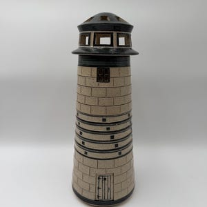 May include: A ceramic lighthouse sculpture featuring a speckled beige body with a brick pattern, dark blue horizontal bands, and a dark blue top section with cut-out windows. The structure includes a small rectangular door and a single window.