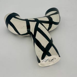 May include: A cream-colored ceramic vase with a unique, curved shape. The vase is decorated with bold black intersecting lines, creating a striking geometric pattern. The vase has a speckled texture and a modern aesthetic.